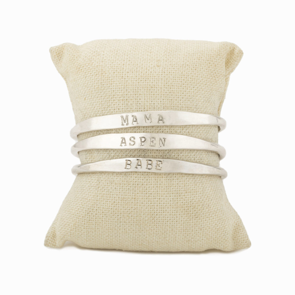 Three silver cuffs on a bracelet pillow with the words "Mama", "Aspen", and "Babe" stamped on each bracelet respectively.