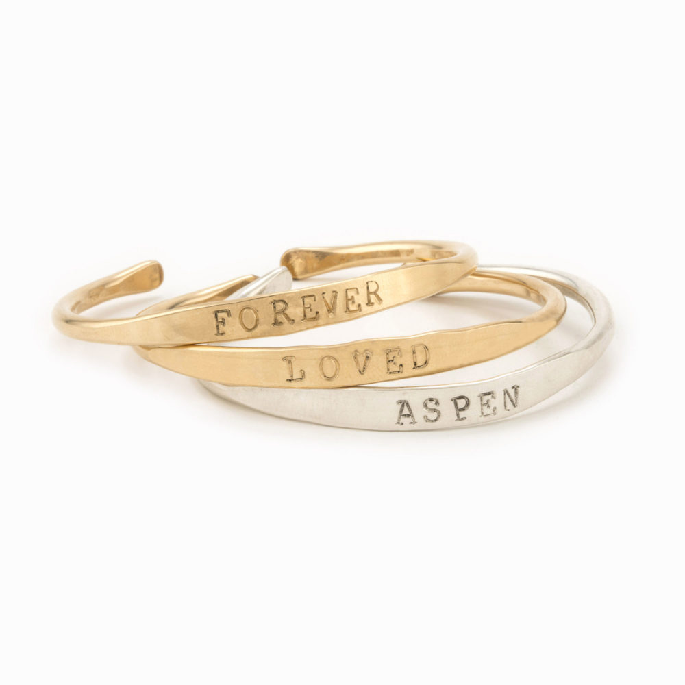 Two 14k gold filled cuffs one with the word "Forever" and one with the word "Loved" stamped into them and stacked on top of a third silver cuff with the word "Aspen" stamped on it.