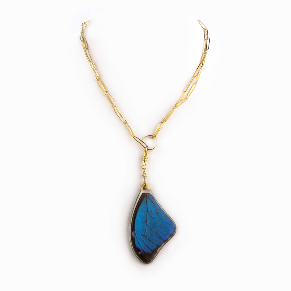 Close up on an electric blue butterfly wing hanging from a thin 14k gold-filled paperclip chain necklace.