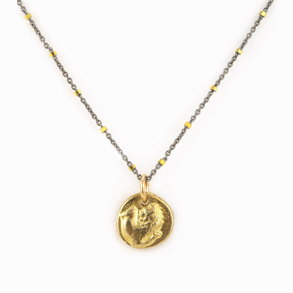 Close up of a stamped bass coin charm hanging forma gold vermeil beeded necklace with oxidized silver chain.
