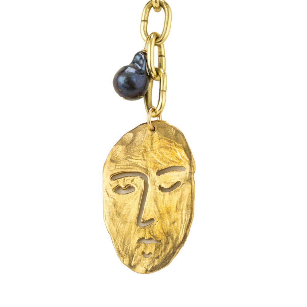 Close up on a brass face charm and tahitian pearl hanging from a thick brass chain.