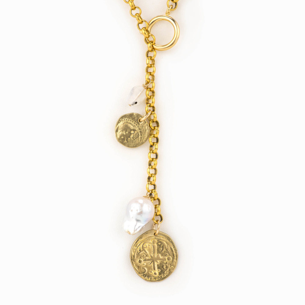Close up on a baroque pearl, small moon stone, and brass antique coin charms hanging from a brass rolo chain necklace.