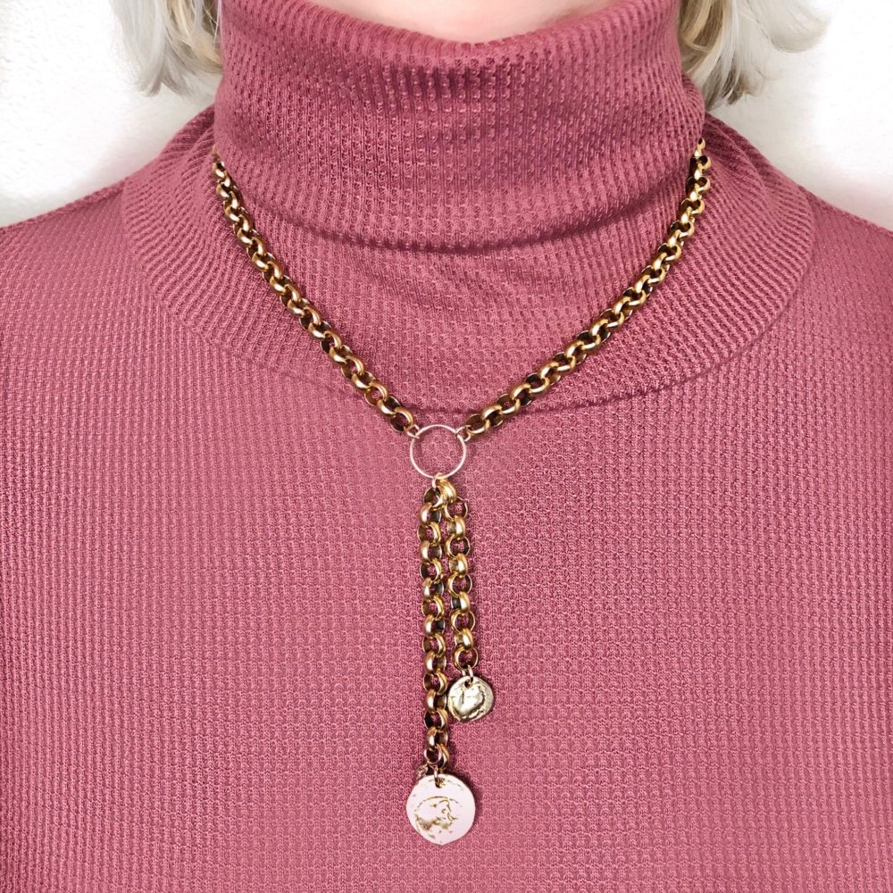 Close up on a woman's torso wearing a brass rolo chain necklace with 2 chain drops, each finished with a brass coin.