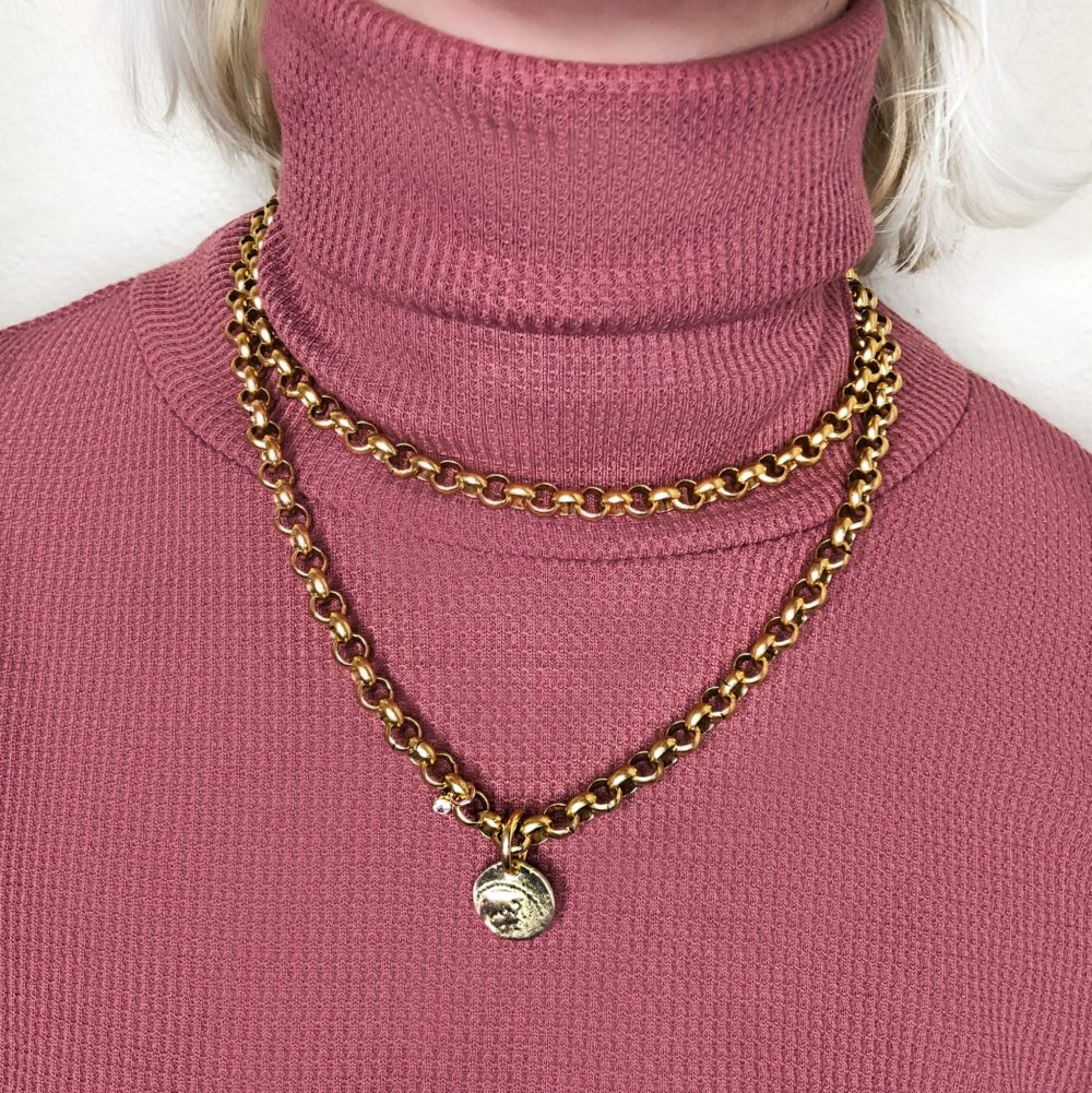 A close up on a woman's neck wearing a layered necklace with brass rolo chain, double-wrapped and antique gold coin over a sweater.