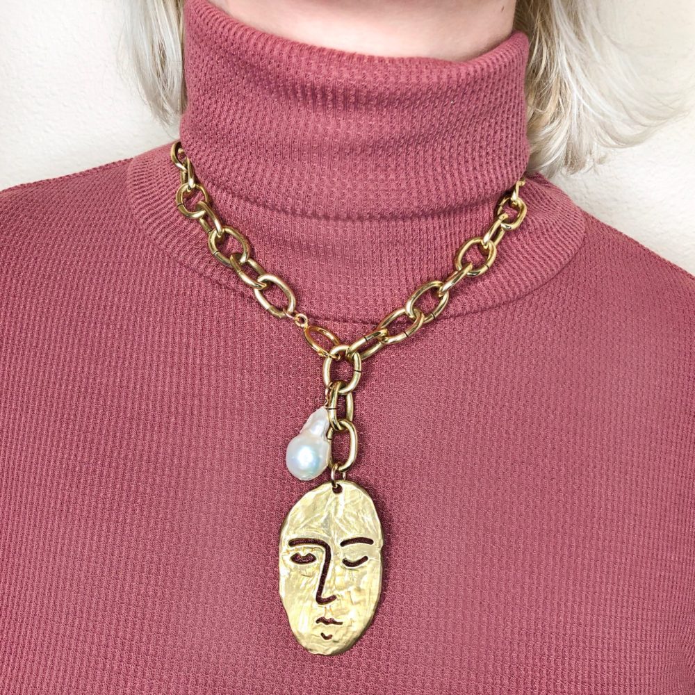 Woman wearing a brass chain necklace with a large carved face charm and baroque pearl.