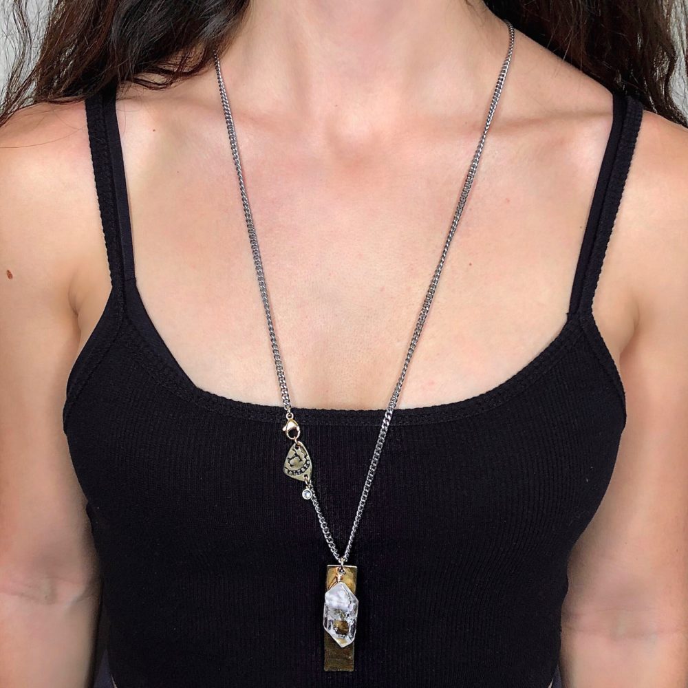 Close up on a woman's neck wearing a flat silver chain necklace with gold tag and drop herkimer diamond quartz charm at full length.