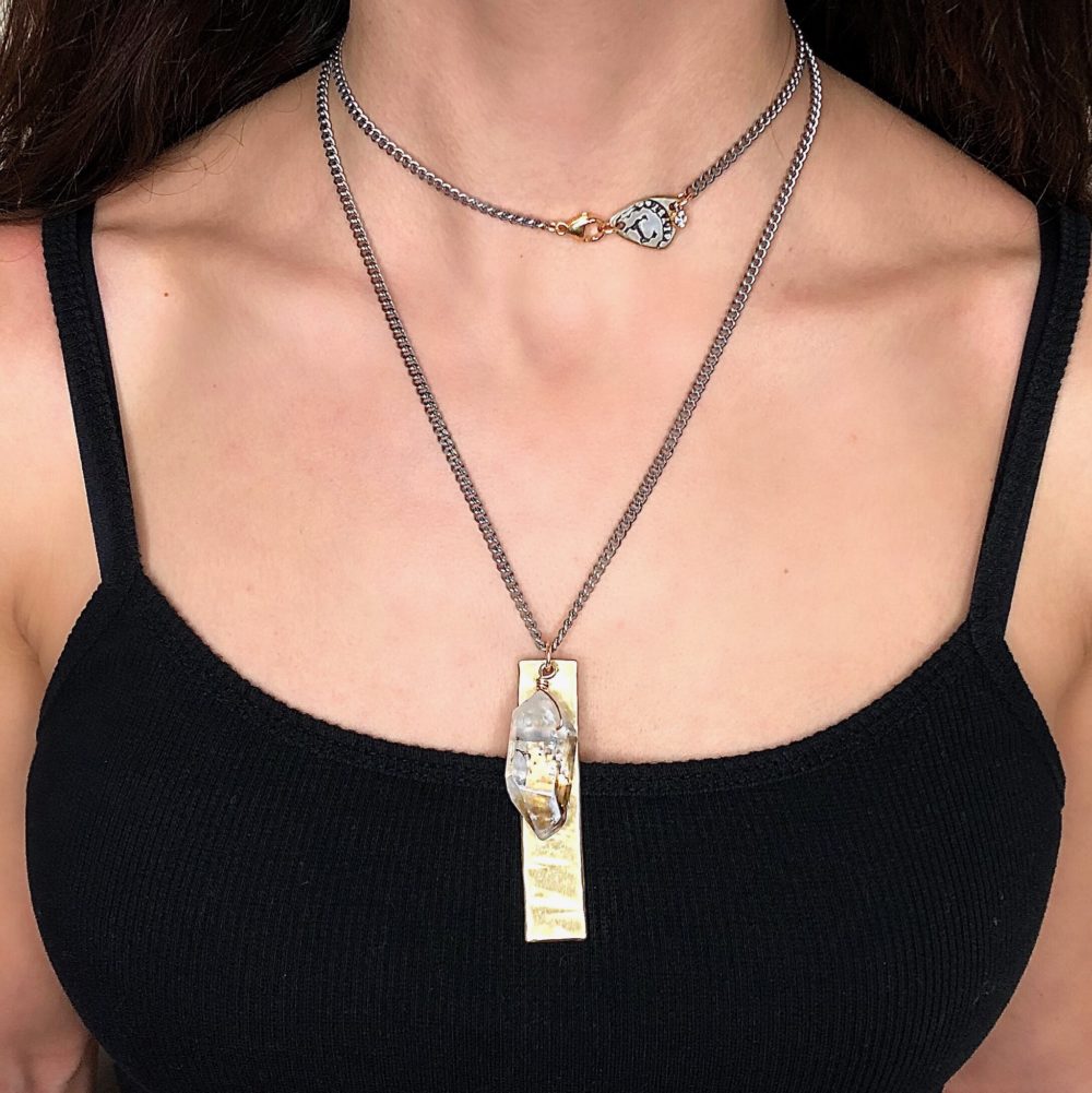 Close up on a woman's neck wearing a flat silver chain necklace with gold tag and drop herkimer diamond quartz charm doubled up.