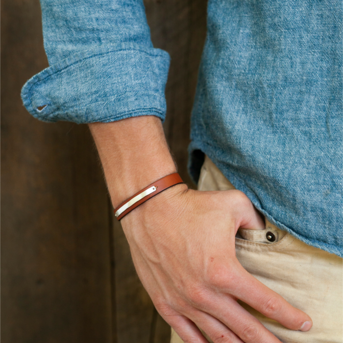 Close up on a man wearing a blue shirt's wrist wearing a brown leather bracelet with sterling silver bar inlay.