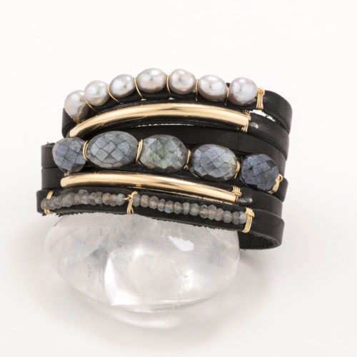A black leather shred bracelet featuring labradorite beads and pearls and finished with a 14k gold bar resting on a chunk of quartz.